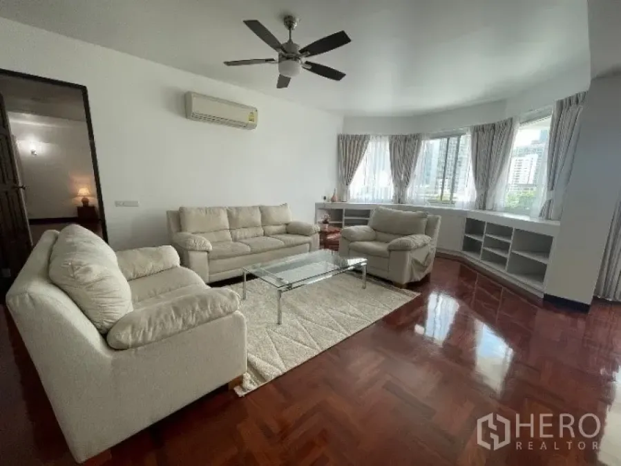 Condominium for rent in Khlong Toei, Bangkok - Spacious living room with parquet wood floors, cream sofas, ceiling fan, bay window, and city views in a Khlong Toei condo.