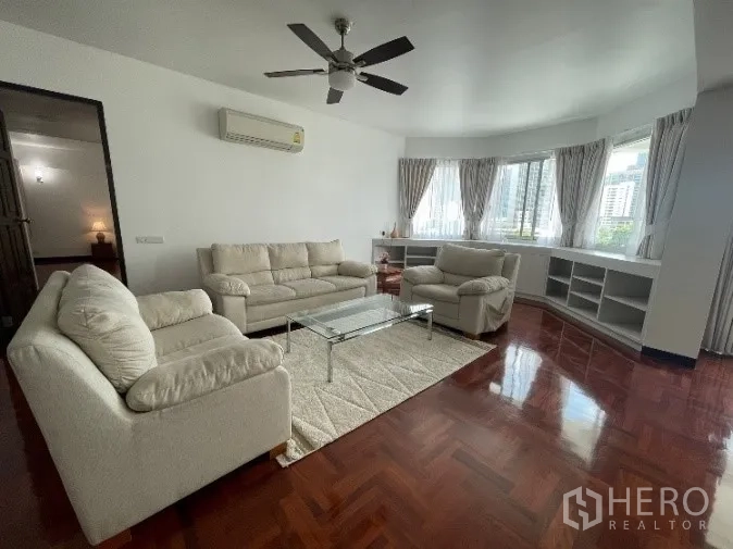 Condominium for rent in Khlong Toei, Bangkok - Spacious living room with parquet wood floors, cream sofas, ceiling fan, bay window, and city views in a Khlong Toei condo.