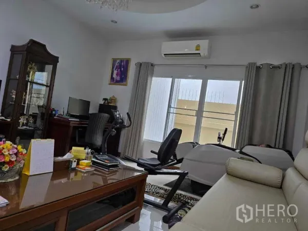 Land for sale - Home office corner with desk, air-conditioning, and a recumbent bike beside large windows.