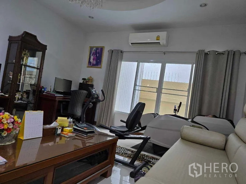 Land for sale - Home office corner with desk, air-conditioning, and a recumbent bike beside large windows.