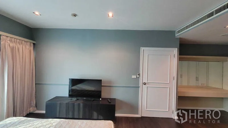 Condominium for rent in Khlong Toei, Bangkok - Master bedroom wall with TV on a low console, curtained glass doors and a white interior door.