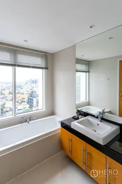 Condominium for rent in Watthana, Bangkok - Bathroom with a bathtub set by a large window overlooking the Bangkok skyline and a wide vanity mirror.
