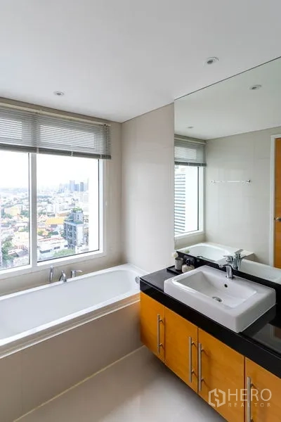 Condominium for rent in Watthana, Bangkok - Bathroom with a bathtub set by a large window overlooking the Bangkok skyline and a wide vanity mirror.