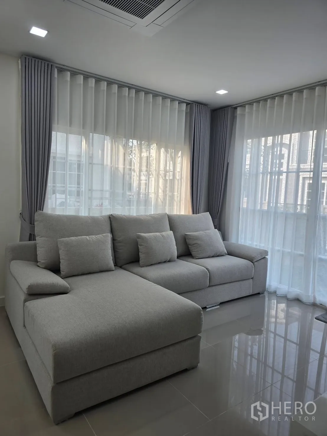 Semi-detached House for rent in Mueang Samut Prakan, Samut Prakan - Corner living area featuring a gray L-shaped sofa, recessed lights and wraparound windows with drapes.