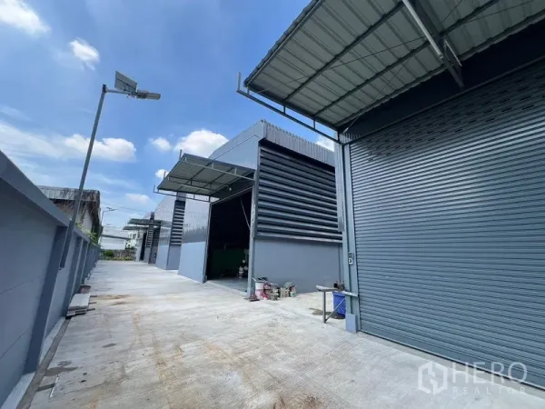 Warehouse for rent in Prawet, Bangkok - Wide concrete driveway in a warehouse compound with roller shutter units and street lighting.