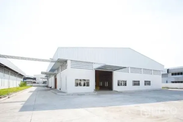 工厂 出租在Bang Pakong, Chachoengsao - 现代白色仓库式厂房正面，高山墙屋顶与大型卷闸门，卡车通行方便。