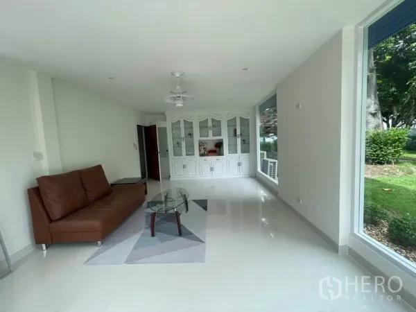Detached House for rent in Khlong Toei, Bangkok - Bright living room with floor-to-ceiling windows, brown sofa, glass coffee table and built-in white cabinet overlooking the garden.