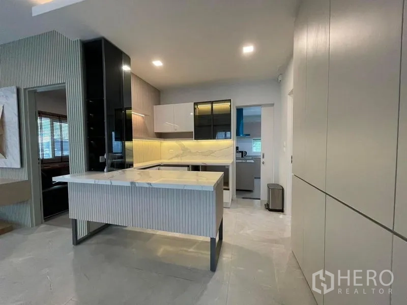 Detached House for rent in Prawet, Bangkok - Stylish show kitchen with marble island, glossy cabinetry and under‑cabinet lighting leading to the Thai kitchen.