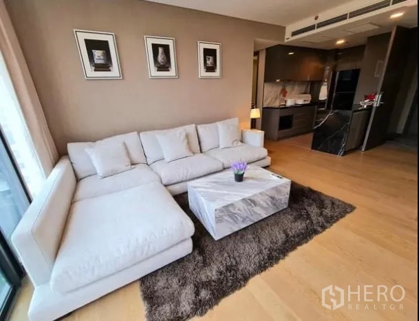 Condominium for rent in Khlong Toei, Bangkok - Warm living room with white L‑shaped sofa, marble coffee table and rug, open to the kitchen.