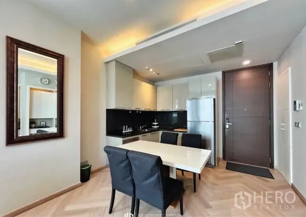 Condominium for rent in Watthana, Bangkok - Compact L-shaped kitchen with black backsplash, dining table for four and entry door.
