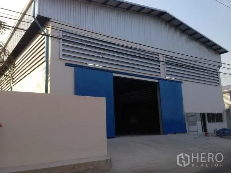 货栈 出租在Mueang Samut Prakan, Samut Prakan - 金属夹芯板外立面仓库，配大幅蓝色推拉门与车道入口。