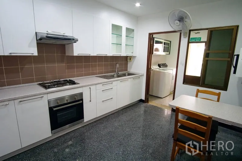 Apartment for rent in Watthana, Bangkok - Modern Western kitchen with white cabinets, gas hob, oven, double sink, and breakfast table leading to a laundry room.
