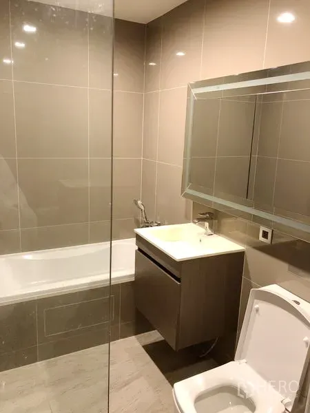 Condominium for rent in Watthana, Bangkok - Modern bathroom with bathtub, vanity, and large mirror in neutral tiles.
