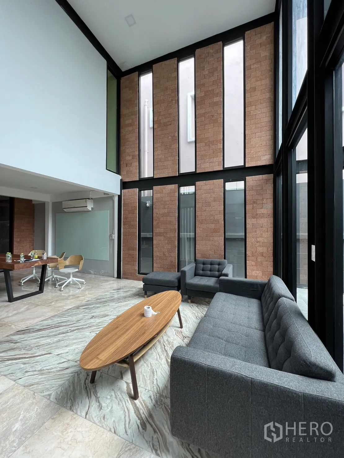 Detached House for rent or sale in Phra Khanong, Bangkok - Double-height living room with brick feature wall, floor-to-ceiling windows, gray sofas and marble-look flooring.
