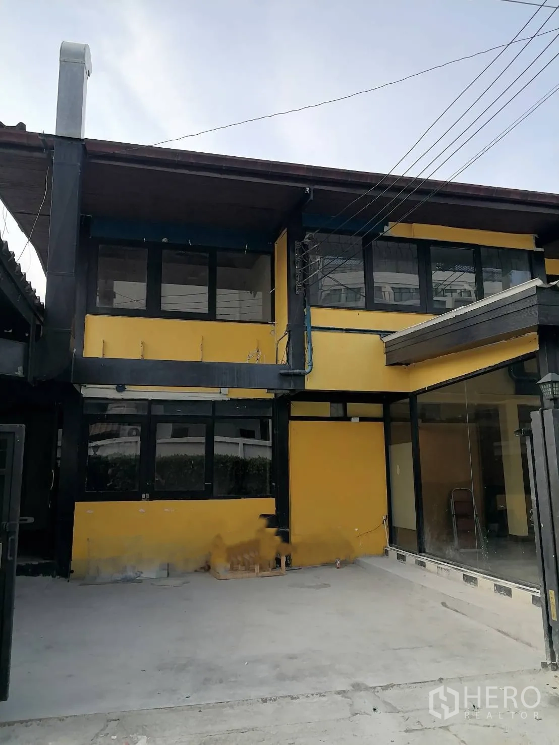 บ้านเดี่ยว for rent in Watthana, Bangkok - บ้านเดี่ยวสองชั้นสีเหลืองกรอบสีดำ มีหน้าต่างบานใหญ่และลานคอนกรีตด้านหน้า.