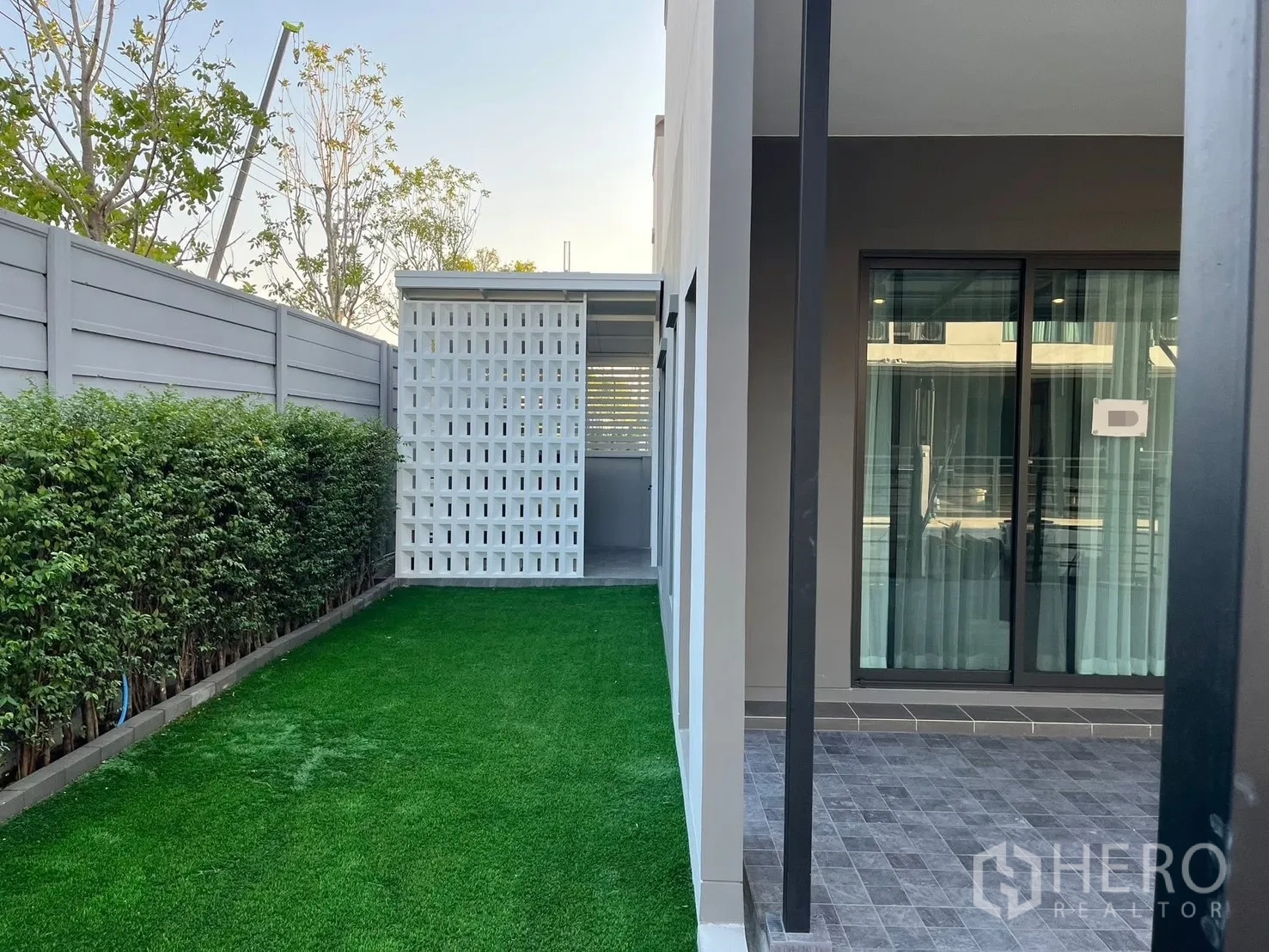 Townhouse for rent in Bang Phli, Samut Prakan - Side garden with artificial grass, hedge, and a covered patio beside the sliding doors of a modern townhouse.