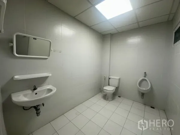 Retail Space for rent - Private tiled restroom with sink, mirror, toilet, and urinal under bright ceiling lighting.