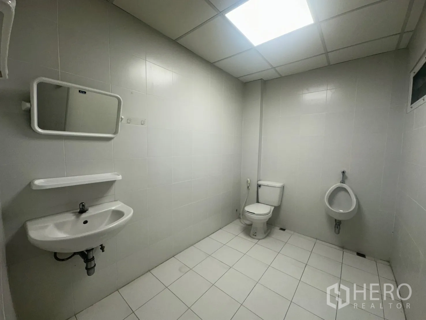 Retail Space for rent - Private tiled restroom with sink, mirror, toilet, and urinal under bright ceiling lighting.