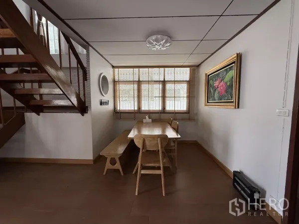 Detached House for rent in Phra Khanong, Bangkok - Dining area beside a wooden staircase with window blinds and framed artwork.