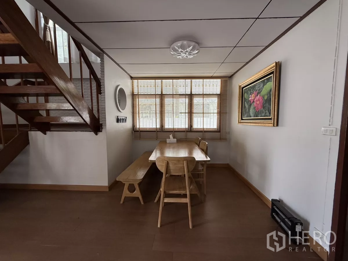 Detached House for rent in Phra Khanong, Bangkok - Dining area beside a wooden staircase with window blinds and framed artwork.