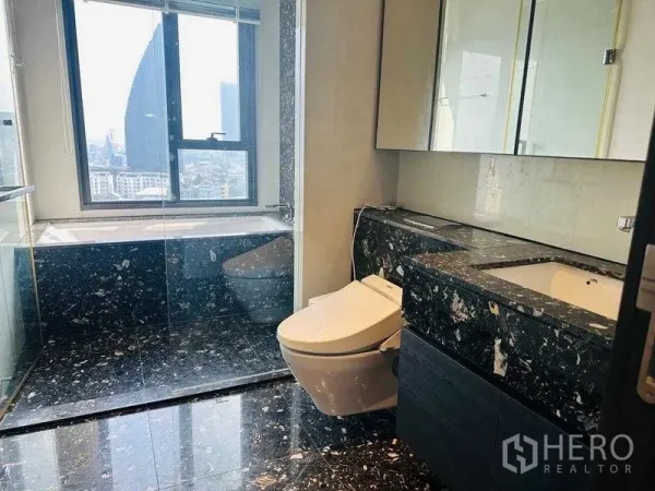 Condominium for rent in Khlong Toei, Bangkok - Modern bathroom with black stone vanity, bathtub by the window, and city view.