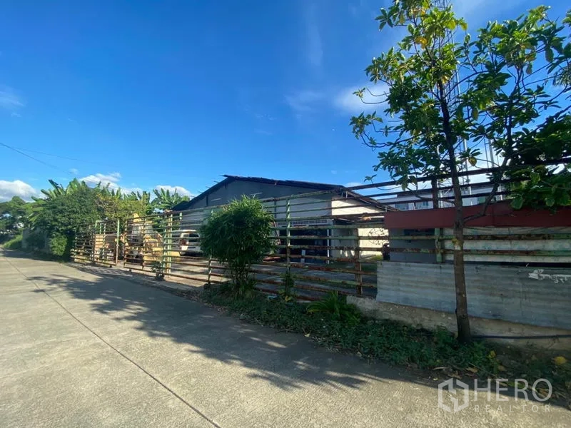 Warehouse for rent in Suan Luang, Bangkok - Street frontage showing long metal fence screening the warehouse with trees along the pavement.