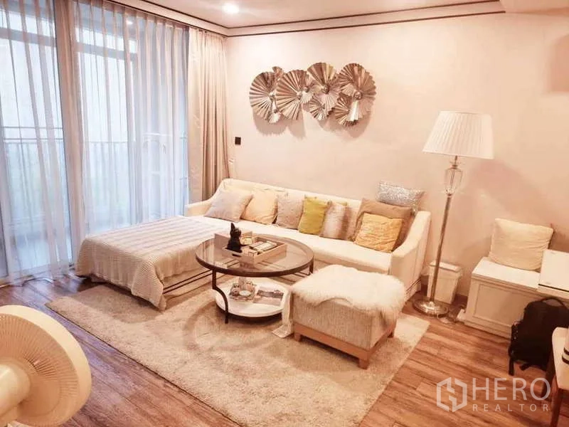Condominium for sale in Bang Rak, Bangkok - Living area with sofa, ottoman, bedside-style lamp and balcony doors framed by long curtains.