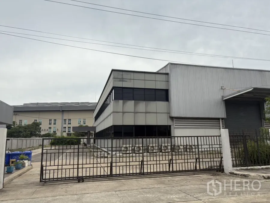 Factory for rent or sale in Samut Prakan - Gated entrance with metal fence facing the office and warehouse facade.
