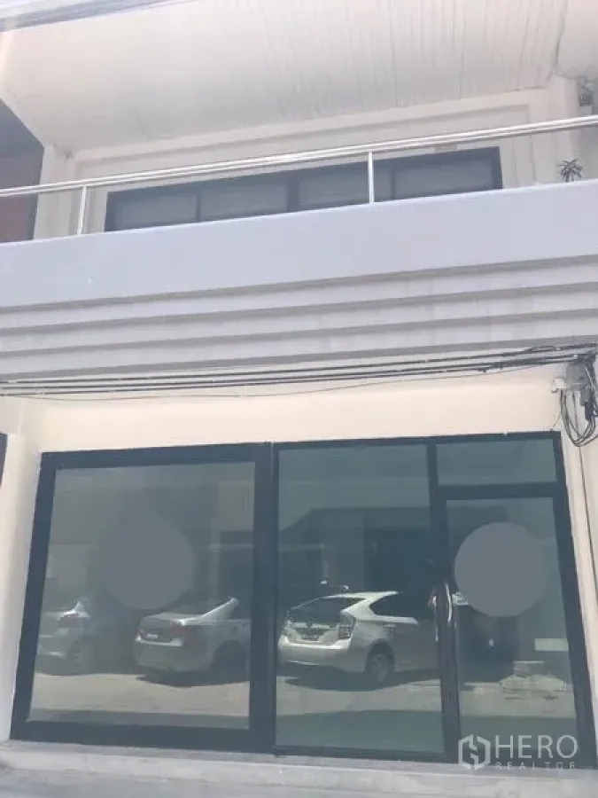 零售空间 出租在Watthana, Bangkok - 一层店面外观，黑色框架落地玻璃滑门，上方有阳台，玻璃倒映停放车辆。