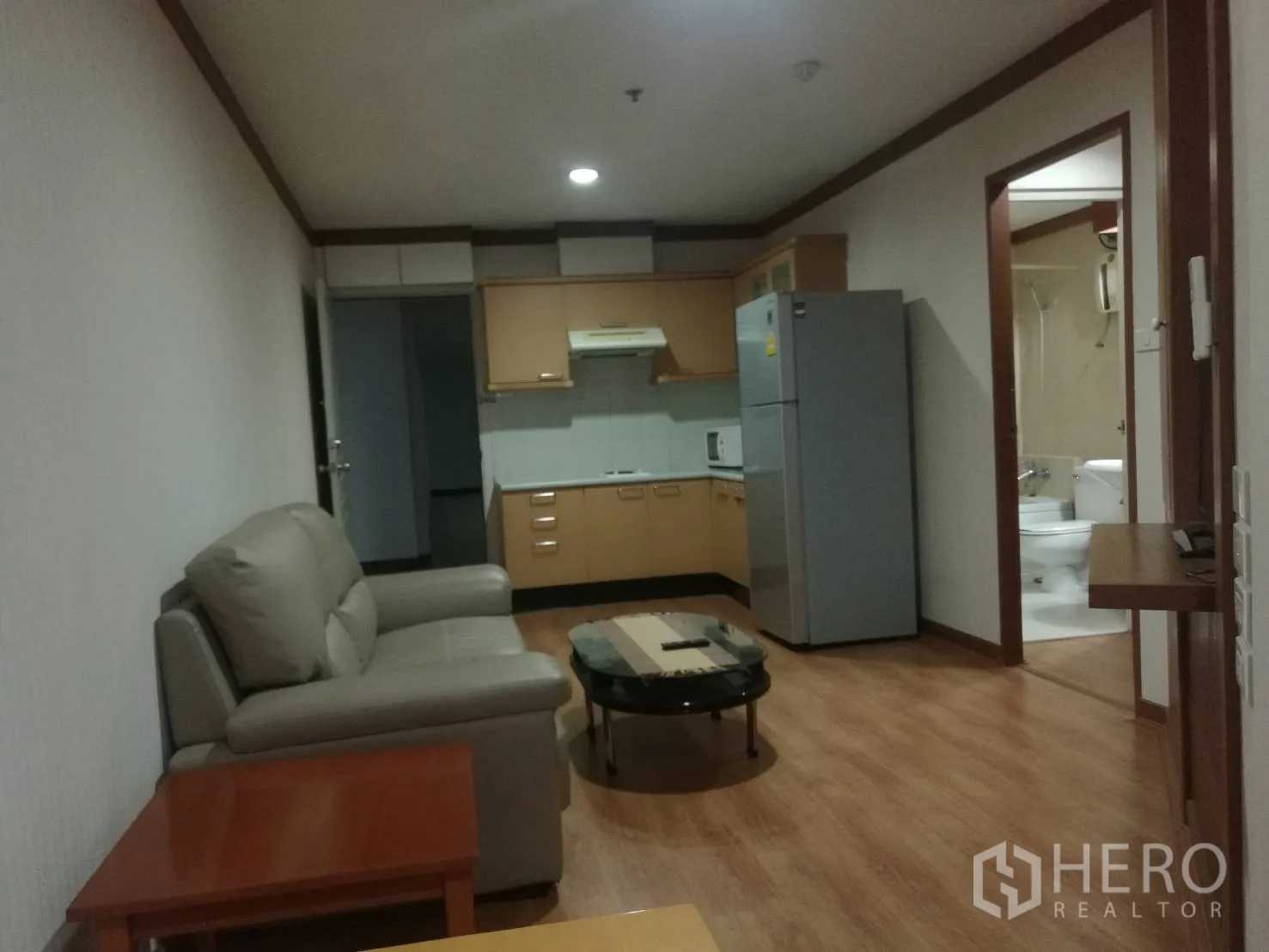 Condominium for rent in Khlong Toei, Bangkok - Open-plan living room with sofa, wood floors and a fitted kitchen with fridge.