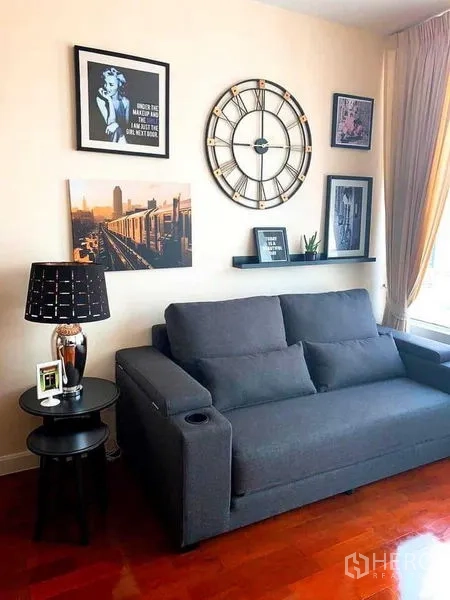 Condominium for rent in Khlong Toei, Bangkok - Living room with gray sofa, large wall clock, framed art, and daylight curtains.