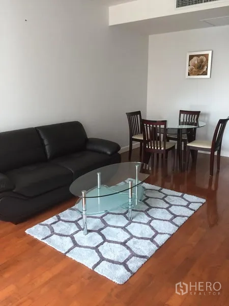 Condominium for rent in Khlong Toei, Bangkok - Open-plan lounge with black leather sofa, glass coffee table, area rug and round dining set on wooden floors.