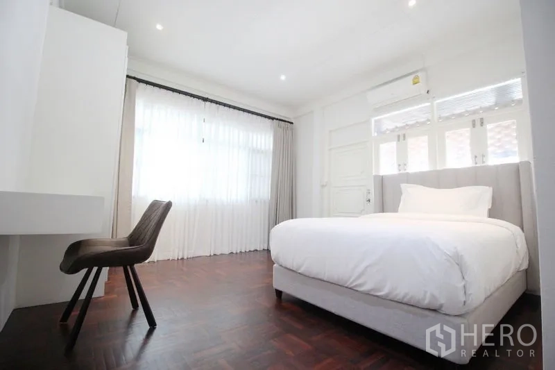 Detached House for rent in Chatuchak, Bangkok - Sunlit bedroom with full‑height window, white bedding, work desk and chair, parquet floor, and air conditioning.