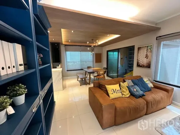 บ้าน ให้เช่าในBang Phli, Samut Prakan - โซนรับแขกและทานข้าวแบบเปิดโล่ง โซฟาหนัง ชั้นบิลท์อินสีน้ำเงิน ฝ้าไม้ และหน้าต่างบานใหญ่