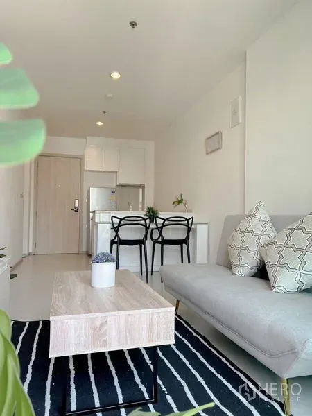 Condominium for rent or sale in Khlong Toei, Bangkok - Bright living room with gray sofa, striped rug, coffee table, two-seat dining and compact white kitchen.