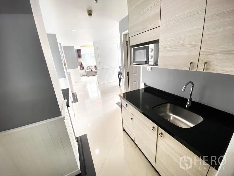 Condominium for rent in Khlong Toei, Bangkok - Galley kitchen with light wood cabinetry, black countertop, sink and microwave.