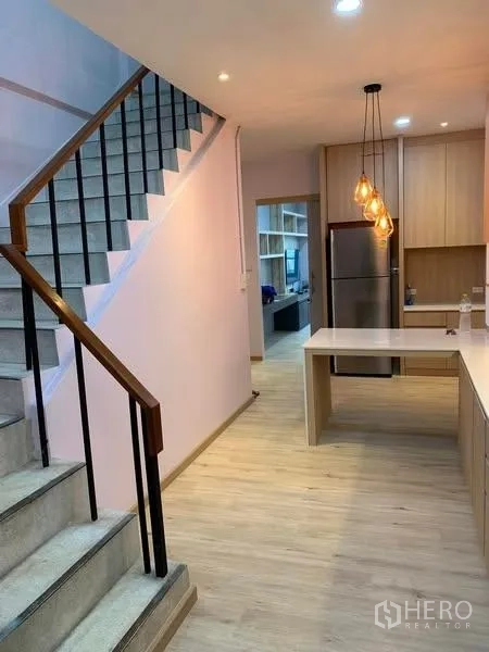 Townhouse for rent - Stair hall with wood handrail leading to an open-plan kitchen and dining area with fridge and warm lighting.