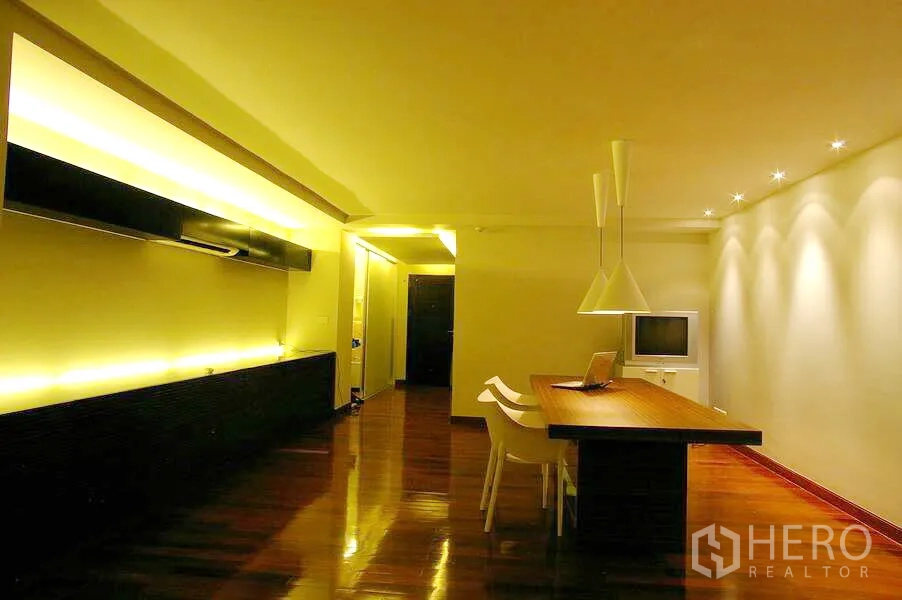 公寓 出租在Watthana, Bangkok - 开放式客餐厅，暖色灯光，长木餐桌、白色椅子，尽头为落地窗与沙发。