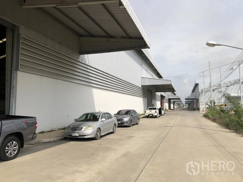 Factory for rent in Wang Noi, Phra Nakhon Si Ayutthaya - Row of warehouse façades with large canopies and roadside parking on a concrete lane.
