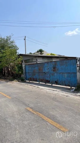 Warehouse for rent in Nong Khaem, Bangkok - Street view of a gated warehouse entrance with overhead power lines and roadside trees in Nong Khaem.