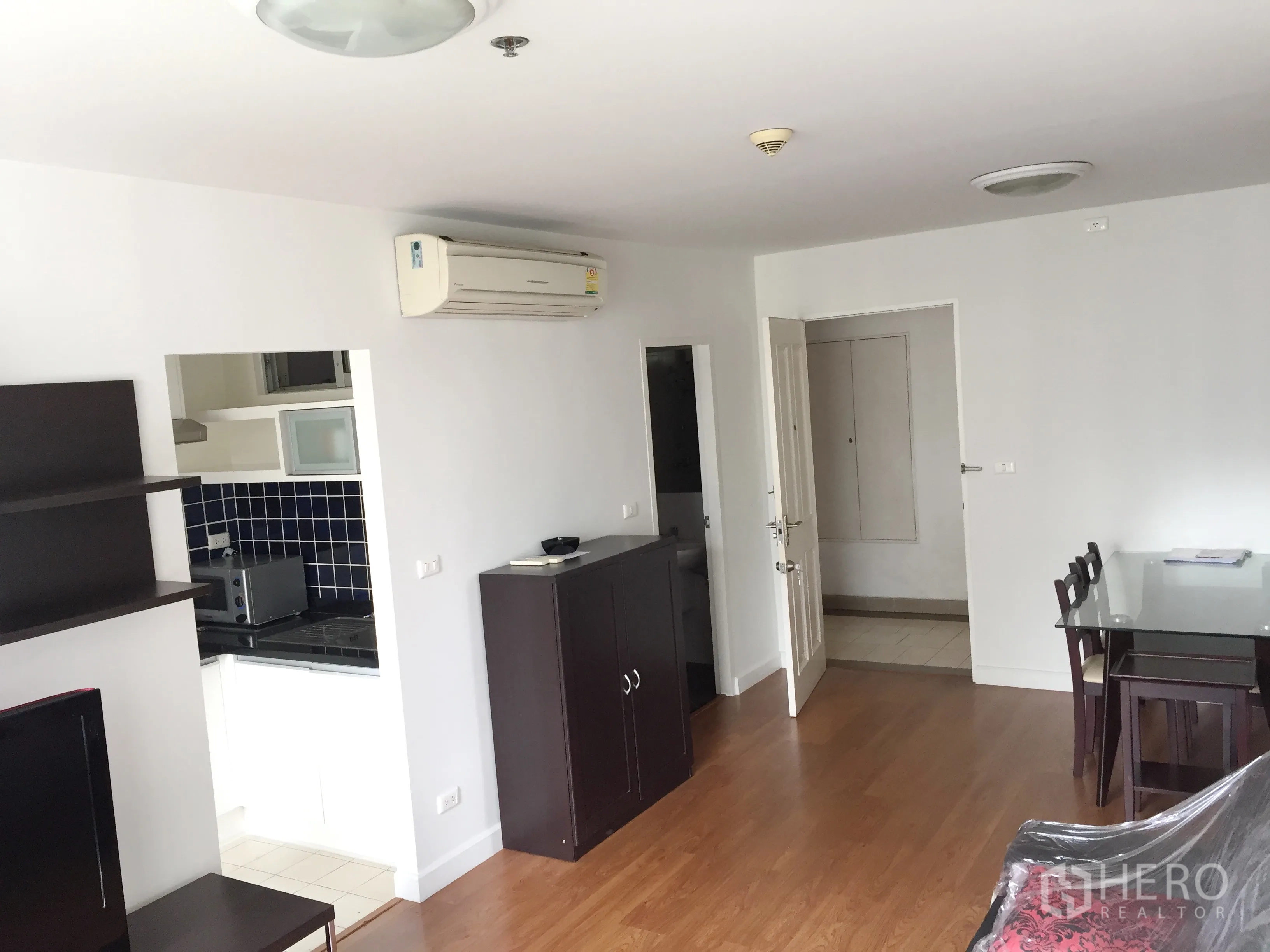 Condominium for rent in Khlong Toei, Bangkok - Open-plan living and dining area with air conditioner, glass dining table and doorway to the kitchen.