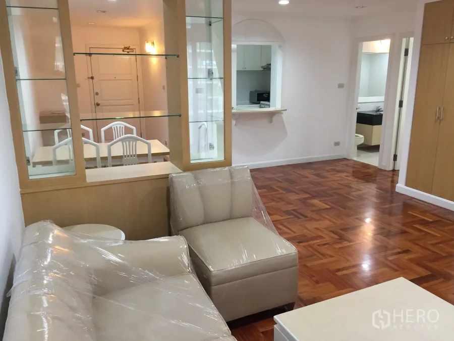 公寓 出租在Watthana, Bangkok - 客厅带玻璃展示隔断与米色沙发，亮面人字形木地板通向玄关与卫浴。
