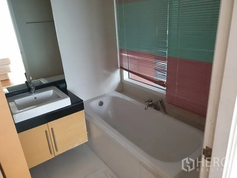 คอนโด ให้เช่าในWatthana, Bangkok - ห้องน้ำพร้อมเคาน์เตอร์อ่างล้างหน้า ถัดจากอ่างอาบน้ำสีขาวและหน้าต่างที่ติดมู่ลี่สีสัน