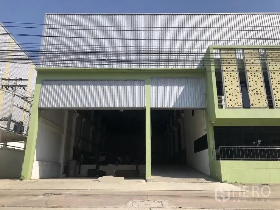 Factory for sale in Bang Phli, Samut Prakan - Street-facing factory exterior with green frame, twin high roller shutter doors, tall metal roof and wide driveway access in Bang Phli.