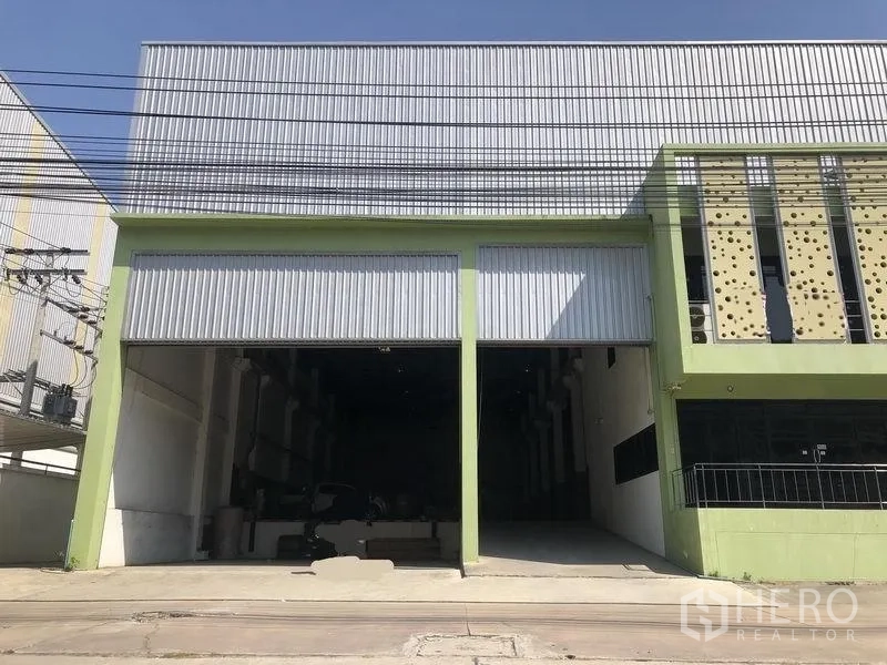 Factory for sale in Bang Phli, Samut Prakan - Street-facing factory exterior with green frame, twin high roller shutter doors, tall metal roof and wide driveway access in Bang Phli.