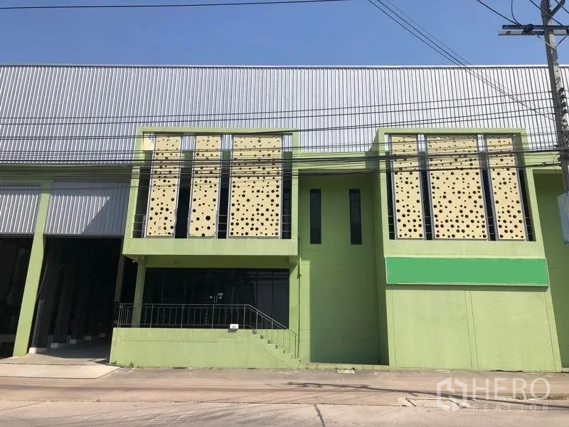Factory for sale in Bang Phli, Samut Prakan - Factory frontage with perforated facade panels, staircase to glass-front office and adjoining high-bay warehouse under clear blue sky.