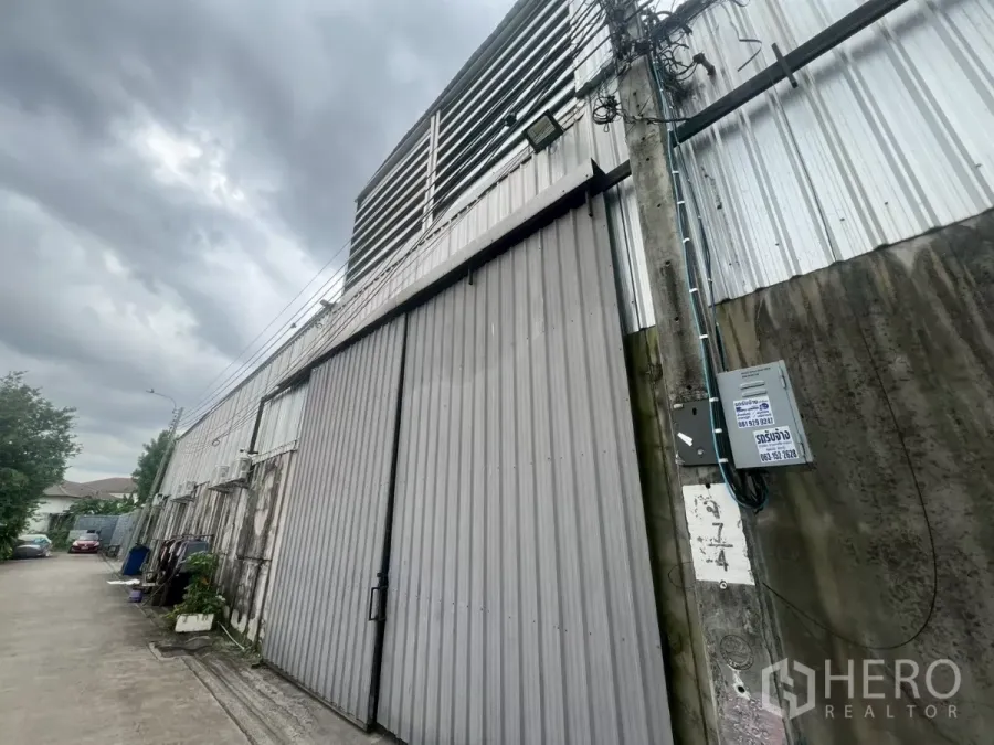 Warehouse for rent - Street-facing corrugated metal warehouse facade with tall sliding gate and utility pole under a cloudy sky.
