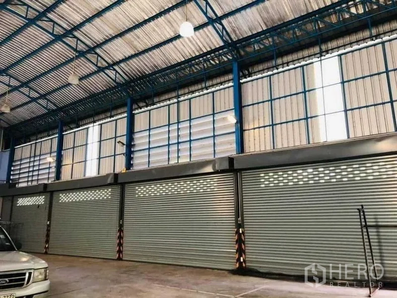 Warehouse for rent in Nakhon Pathom - Row of metal roller shutters beneath tall windows and blue steel trusses inside the warehouse.
