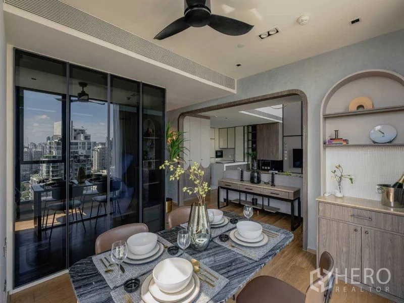 Condominium for sale in Khlong Toei, Bangkok - Open-plan dining area beside glass sliding doors and kitchen, with city views and a ceiling fan.
