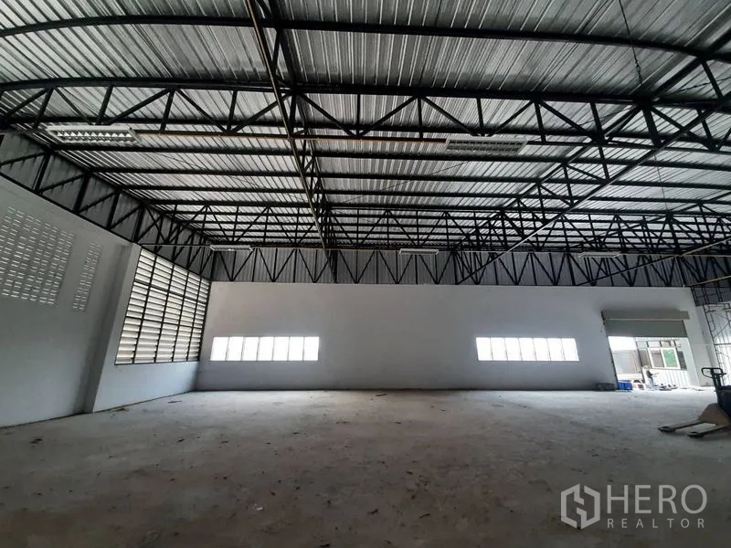 Warehouse for rent in Lam Luk Ka, Pathum Thani - Open clear-span storage area with metal truss ceiling, side windows, and a roller shutter entry.